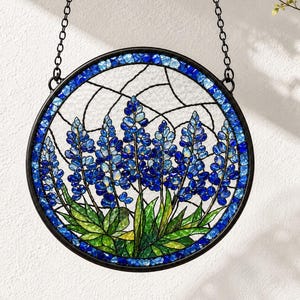 Peut inclure: Un capteur de soleil circulaire en vitrail représentant des fleurs de lupin bleues avec des feuilles vertes. Le motif est encadré par un contour en métal sombre et suspendu par une chaîne noire, mettant en valeur des textures de verre variées.