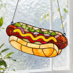May include: A stained glass suncatcher shaped like a hot dog, featuring a tan bun, a brown sausage, a wavy yellow mustard line, and green and purple relish accents. The piece is suspended by two thin metal chains attached to the top edges.