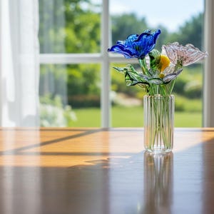 May include: A glass vase with a bouquet of colorful glass flowers sits on a wooden table. The flowers include blue, pink, and yellow petals with green leaves. The vase is clear with vertical lines. A window and white curtains are in the background.