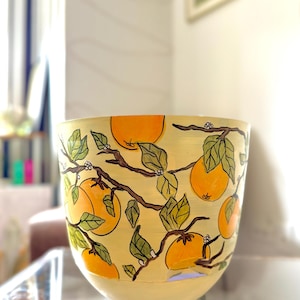 Puede incluir: Un cuenco de cerámica pintado a mano con fondo amarillo claro. Decorado con naranjas, hojas verdes y ramas marrones. El diseño envuelve todo el cuenco, creando una estética vibrante y natural. Ideal para decoración o servir alimentos.