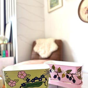 May include: Two rectangular planters, one light green and one pink, with white interiors. The green planter features pink and blue floral designs, while the pink planter has a blackberry and white flower motif. Both sit on matching trays.