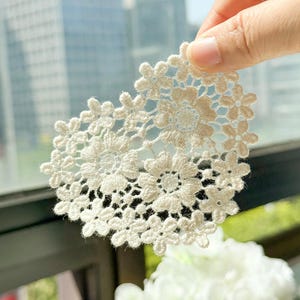 May include: Close-up of a cream-colored, floral lace applique. The delicate lace features a repeating pattern of flowers and leaves, creating an intricate design. The applique is held up against a blurred background, highlighting the texture and detail of the lace.