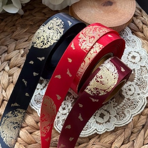 May include: Three spools of decorative ribbon in black, red, and burgundy. Each ribbon features a gold dragon design and cloud accents. The ribbons are displayed on a woven surface with a lace doily.