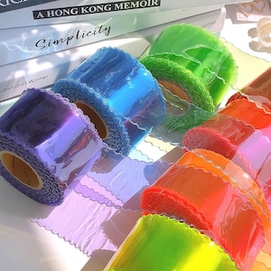 May include: A collection of colorful, translucent rolls of decorative ribbon. The ribbons are in shades of purple, blue, green, orange, and pink. Each roll has a scalloped edge, and the light reflects off the shiny surface.