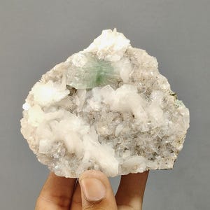 May include: A rough, natural mineral specimen featuring a cluster of white crystal formations with a central, light green crystal. The mineral is held in a hand against a gray background.