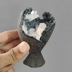 Könnte beinhalten: Eine kelchförmige Geodenskulptur mit einer rauen, dunkelgrauen Außenseite und einem hohlen Inneren, das mit funkelnden dunkelgrauen Kristallen und markanten, durchscheinenden weißen Kristallformationen in der Mitte ausgekleidet ist.