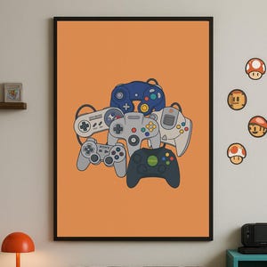 May include: A framed art print featuring a collection of retro video game controllers in various colors, including blue, gray, and black, set against an orange background. The controllers are stacked together, showcasing different designs.