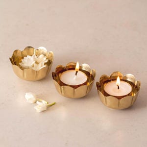 May include: Three gold-colored lotus-shaped candle holders. Two holders contain lit white candles, while the third holds white flowers. The holders are arranged on a light-colored surface, creating a warm and inviting ambiance.