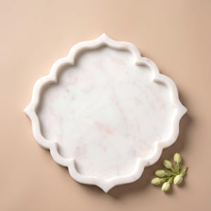 May include: A white marble tray with a scalloped edge, featuring subtle pink veining. The tray has a quatrefoil shape and is set against a neutral background. A small sprig of white flower buds is placed on the side.