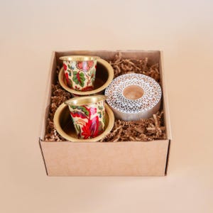 May include: A gift set in a cardboard box featuring two small, gold-rimmed cups with floral patterns in red, green, and yellow. A decorative, circular candle holder with a carved design is also included.