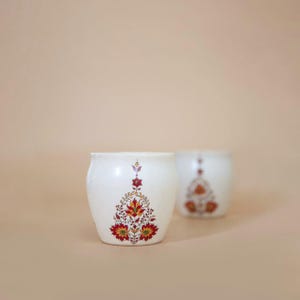 May include: Two white ceramic pots with a floral design in red, yellow, and green. The pots have a rounded shape and are set against a neutral background. The floral pattern is symmetrical and detailed.