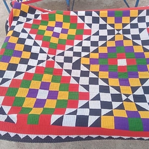 May include: A colorful, geometric quilt with a checkerboard pattern in red, yellow, green, purple, black, and white. The quilt has a red border with a black and white zigzag trim. The design is symmetrical and eye-catching.
