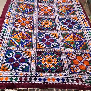 Pode incluir: Um têxtil colorido e estampado com uma borda bordô. O design apresenta formas geométricas em laranja, azul, verde e branco, dispostas em uma grade. O têxtil é provavelmente um item decorativo, como um cobertor ou tapete.