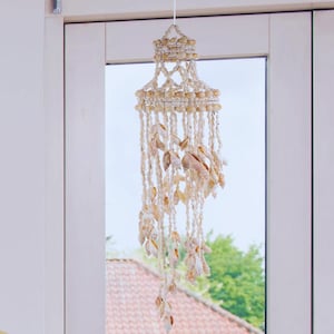 Peut inclure: Mobile décoratif en coquillages, avec un design à plusieurs niveaux et de multiples brins de coquillages. Le mobile est suspendu par une fine ficelle blanche. Les coquillages sont dans des tons beige et crème. En arrière-plan, une fenêtre avec vue sur une maison et des arbres.