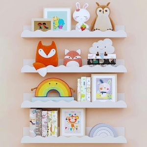Peut inclure: Des étagères blanches en forme de nuages présentent des décorations pour enfants. On y trouve des cadres, des animaux en peluche comme un lapin et un hibou, un arc-en-ciel et des coussins en forme de renard. Des livres et un arc-en-ciel en bois sont également visibles.