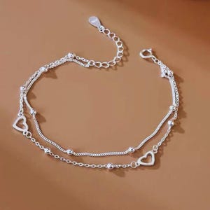 Puede incluir: Una tobillera plateada de doble capa con dijes en forma de corazón y pequeños detalles de cuentas. La tobillera presenta un diseño de cadena delicado y un cierre seguro.