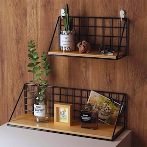 May include: Two-tier wall shelves with a black metal grid design and wooden shelves. The shelves are decorated with a small cactus, a figurine, a framed picture, a glass vase with greenery, and a perfume bottle.