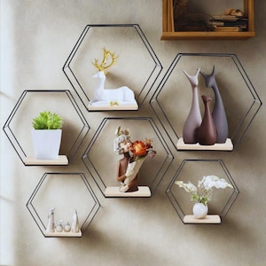 Peut inclure: Ensemble de six étagères hexagonales en métal noir avec des plateformes en bois clair. Chaque étagère présente des objets décoratifs, dont une figurine de cerf blanche, une plante en pot et des vases en céramique. Les étagères sont fixées sur un mur de couleur neutre.