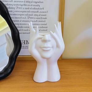 Pode incluir: Um vaso de cerâmica branca em forma de rosto, com as mãos cobrindo os olhos. O vaso tem uma boca sorridente e está colocado sobre uma superfície de madeira. É um item decorativo.
