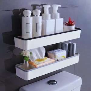 Puede incluir: Estantería de baño blanca y gris de dos niveles. El estante superior contiene botellas blancas y una pequeña planta en maceta. El estante inferior contiene paquetes de pañuelos y rollos de papel higiénico.