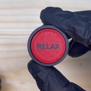 Puede incluir: Un objeto circular con un borde exterior negro y un centro rojo texturizado. La palabra RELAX está grabada en letras mayúsculas negras en el centro de la superficie roja. El objeto tiene una apariencia impresa en 3D con líneas visibles.