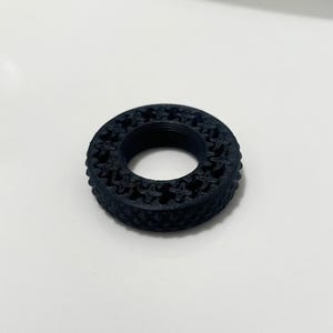 Könnte beinhalten: Ein schwarzes, kreisförmiges Objekt mit gemusterter Außenseite. Das Objekt hat eine Reihe sich wiederholender sternförmiger Formen an seinem äußeren Rand. Das Objekt besteht aus einem festen Material und liegt auf einer weißen Oberfläche.