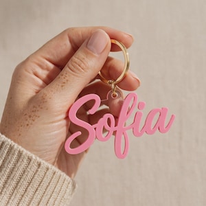 May include: A pink acrylic keychain featuring the name Sofia in a cursive script font, attached to a gold-colored metal split ring. The keychain is made of a smooth, solid pink material and displays the name clearly in a stylized, flowing typeface.