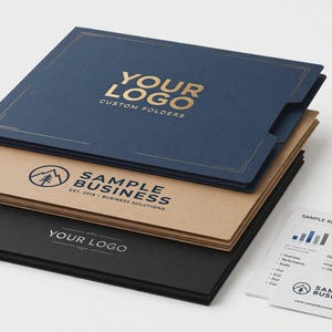 May include: A stack of three rectangular presentation folders in navy blue, tan, and black, featuring gold and black printed logos and text. Beside the folders lies a white document titled Sample Report Q4 2024 with contact information.