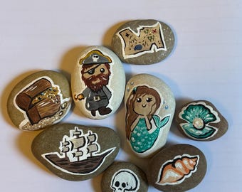 Può includere: Una collezione di sassi dipinti a mano con temi pirateschi e nautici. I disegni includono un pirata, un forziere, una mappa, una nave, una sirena, un teschio, una conchiglia e una perla in una conchiglia. I sassi sono dipinti con colori vivaci.