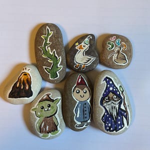May include: A collection of hand-painted rocks featuring whimsical designs. The artwork includes a volcano, a green plant, a duck wearing a party hat, a bag of coins, a goblin, a gnome, and a wizard with a staff. The rocks are painted with bright colors.