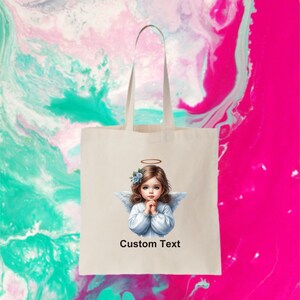 Pode incluir: Uma bolsa de tecido branca com uma imagem impressa de um anjo com uma aur&eacute;ola e flores azuis no cabelo. O anjo est&aacute; vestindo um vestido branco e tem as m&atilde;os juntas em ora&ccedil;&atilde;o. O texto "Custom Text" est&aacute; impresso abaixo da imagem.