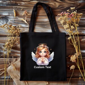 May include: Black tote bag with a white angel illustration and the text "Custom Text". The angel has blonde hair, a halo, and is wearing a white dress. The background is a wooden surface.