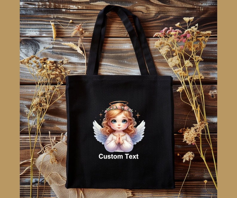 May include: Black tote bag with a white angel illustration and the text "Custom Text". The angel has blonde hair, a halo, and is wearing a white dress. The background is a wooden surface.