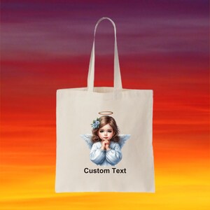 Pode incluir: Uma bolsa tote branca com uma imagem de um anjo com uma aur&eacute;ola e flores azuis no cabelo. O anjo est&aacute; vestindo um vestido branco e tem as m&atilde;os juntas em ora&ccedil;&atilde;o. O texto "Custom Text" est&aacute; impresso abaixo da imagem.