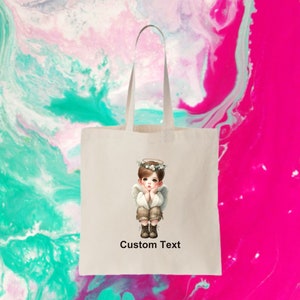 May include: A white tote bag with a design of a young angel with a halo and wings. The angel is wearing a brown shirt and pants and has a light blue halo. The text "Custom Text" is printed below the angel.