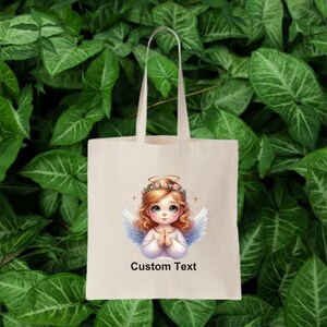 May include: A white tote bag with a design of a cartoon angel with blonde hair and a halo. The angel is wearing a white dress and has her hands clasped in prayer. The text "Custom Text" is printed below the image.