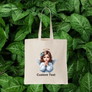 Pode incluir: Uma bolsa tote branca com uma imagem impressa de um anjo com uma aur&eacute;ola e flores azuis no cabelo. O anjo est&aacute; vestindo um vestido branco e tem as m&atilde;os juntas em ora&ccedil;&atilde;o. O texto "Custom Text" est&aacute; impresso abaixo da imagem.