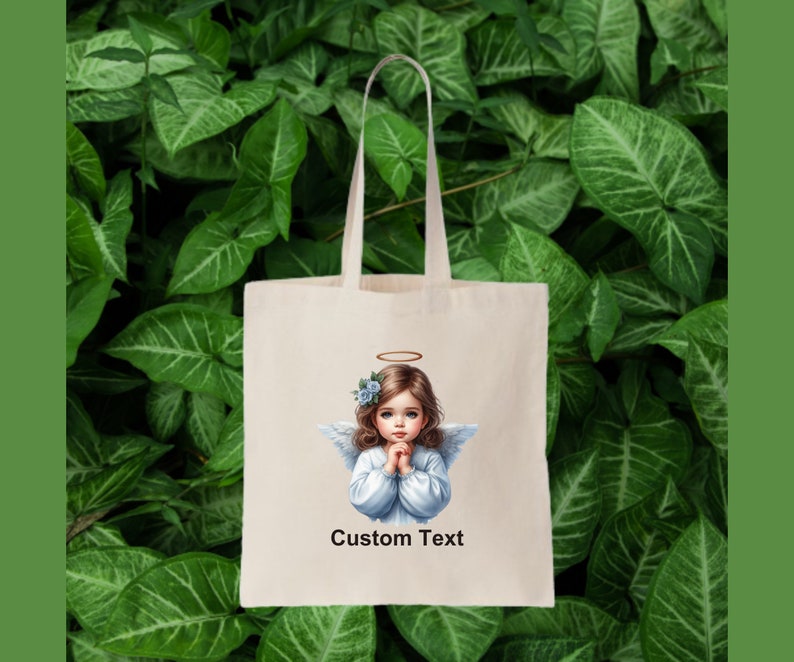 Pode incluir: Uma bolsa tote branca com uma imagem impressa de um anjo com uma aur&eacute;ola e flores azuis no cabelo. O anjo est&aacute; vestindo um vestido branco e tem as m&atilde;os juntas em ora&ccedil;&atilde;o. O texto "Custom Text" est&aacute; impresso abaixo da imagem.
