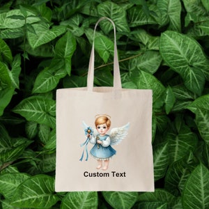 以下が含まれることがあります： 白いトートバッグに、青と白の天使のイラストと「Custom Text」の文字が書かれています。