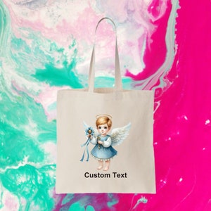 以下が含まれることがあります： 白いトートバッグに、青と白の天使のイラストと「Custom Text」の文字が書かれています。