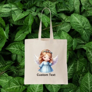 Puede incluir: Una bolsa de tela blanca con un dise&ntilde;o de una ni&ntilde;a &aacute;ngel con cabello rubio y alas azules. El texto "Custom Text" est&aacute; impreso en la bolsa.