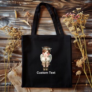 May include: Black canvas tote bag with a white angel illustration and the text "Custom Text".