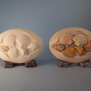 May include: Two oval-shaped ceramic plaques with raised seashell designs. One plaque features a white seashell design, while the other features a colorful seashell design with shades of orange, yellow, and pink.