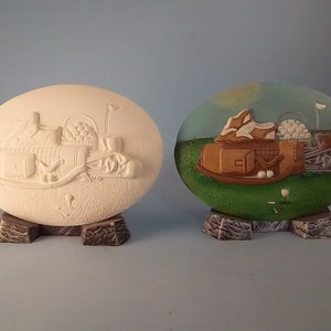 May include: A white ceramic golf-themed oval plaque with a 3D design of golf clubs, balls, and a golf bag. The plaque is on a brown stand. The second plaque is painted with a similar design, but with a green grass background and a blue sky.