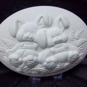 May include: White ceramic oval plaque with a raised design of two bunnies wearing overalls sitting in a field of grass.