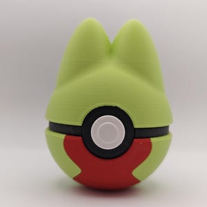 Pode incluir: Um item de novidade impresso em 3D, verde e vermelho, que se assemelha à cabeça de um personagem de desenho animado. O item tem uma faixa preta ao redor do meio com um círculo branco no centro. A parte superior tem duas protuberâncias verdes arredondadas.