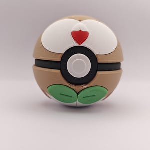 Könnte beinhalten: Eine 3D-gedruckte Pokéball-Replik mit braunem und weißem Design, einem schwarzen Band und einem weißen Mittelkreis. Der Ball hat grüne, blattartige Details und einen roten herzförmigen Akzent.