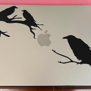 May include: A silver laptop with a black raven silhouette design. Two birds perch on a branch on the upper left, and a single raven sits on a branch on the lower right. The Apple logo is visible.