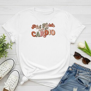 Puede incluir: Una camiseta blanca de manga corta con el texto Plot Twists ARE MY CARDIO en una fuente retro multicolor. La camiseta se muestra junto a unos vaqueros azules, zapatillas de lona blancas, una pequeña planta verde y gafas de sol marrones.