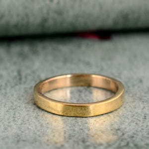 May include: A gold ring with a flat band. The ring has a simple design and appears to be made of metal. The ring is displayed on a textured, grey surface.